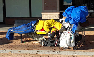 Homeless person sleeping on bench at a shopping center.